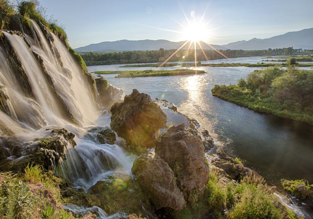 Waterfall cascading into the Snake River at sunrise with sunburst over forested landscape in eastern Idaho