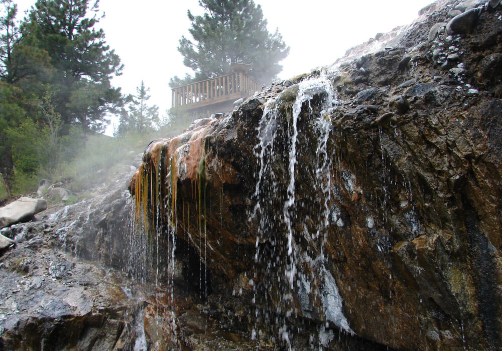 Close-up view of mineral-streaked rock with hot spring water cascading down at Kirkham Hot Springs in Idaho with mist and trees in the background